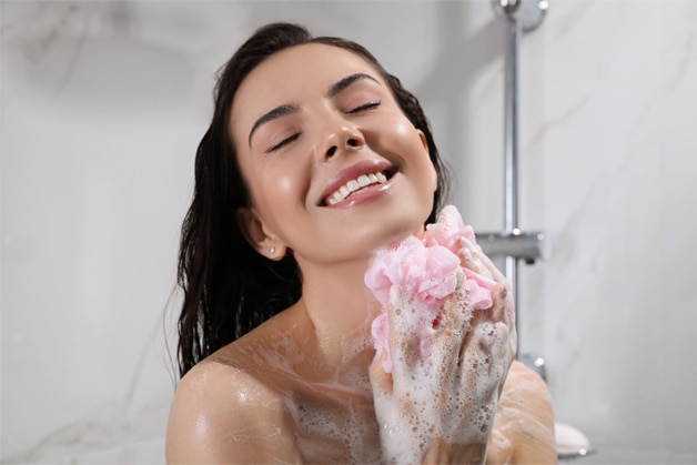 Uma mulher sorridente desfrutando de um banho relaxante enquanto aplica o sérum corporal com uma esponja
