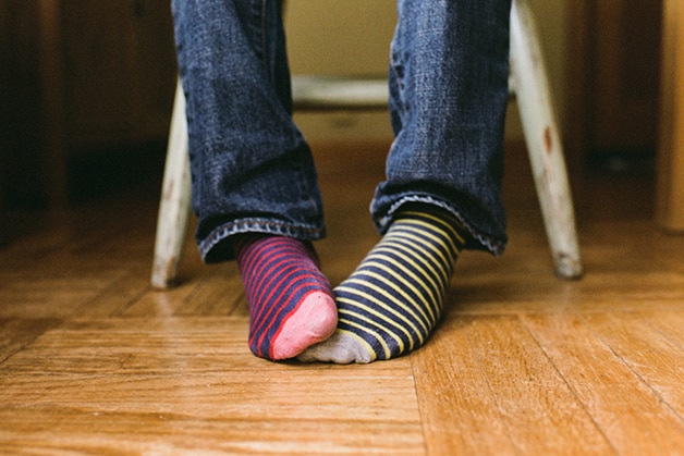 Pessoa usando meias listradas diferentes, uma vermelha e outra azul, representando a transpiração e acúmulo de bactérias que podem causar o chulé