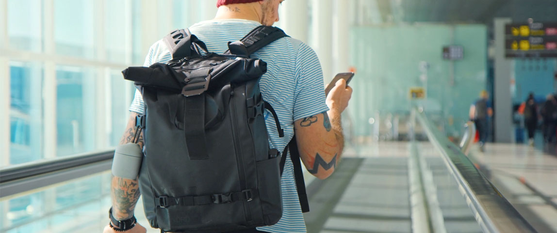 Homem andando em aeroporto com celular na mão e mala de viagem nas costas