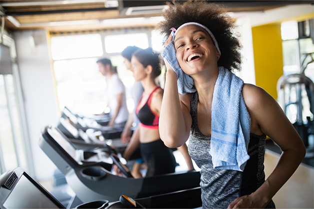 Mulher sorridente na academia após exercício, enxugando o suor com toalha, se preparando para o banho pós o treino.