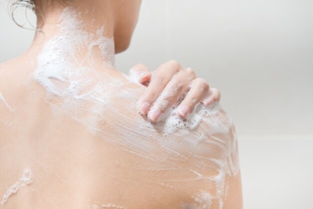 Mulher ensaboando o ombro no banho com espuma suave, usando sabonete para pele com dermatite.
