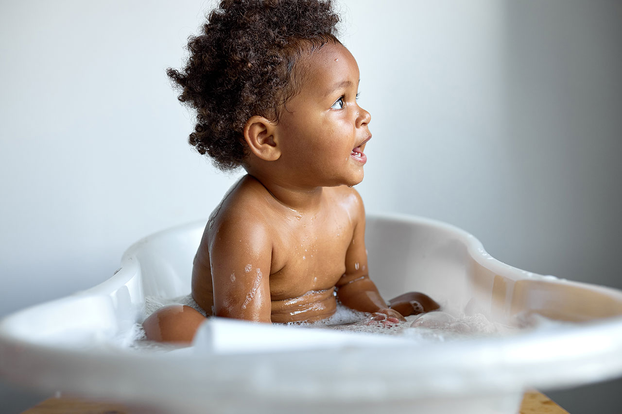 Criança pequena em banheira branca, com espuma e corpo molhado, olhando para o lado em um momento de banho em casa, com fundo desfocado.