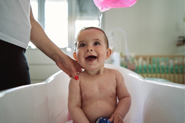 No banho para bebê, adulto despeja água na cabeça da criança com uma caneca para enxaguar o sabonete  líquido Protex.