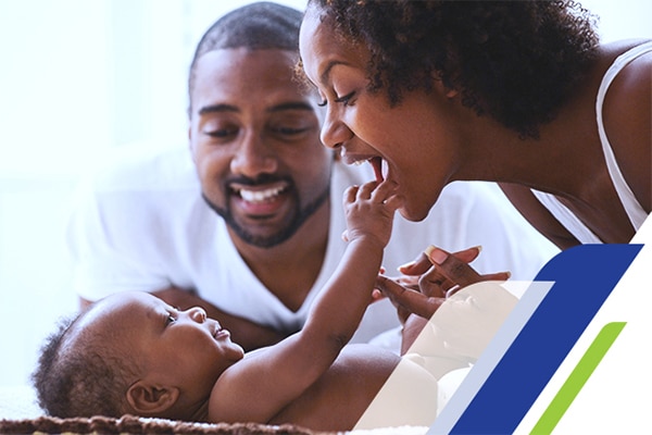 Um bebê deitado tocando com uma mão o rosto da sua mãe, que está sorrindo enquanto seu pai o observa