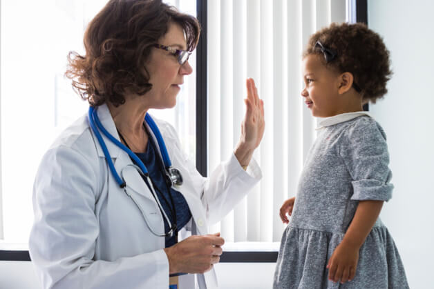 Imagem de uma médica do lado esquerdo e uma menina de 2 anos em pé em cima da cama do lado direito.
